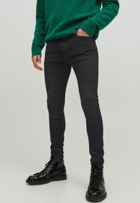 Black slim-fit jeans with a smooth texture, paired with black lace-up boots, and a green sweater. Simple design with no visible embellishments.