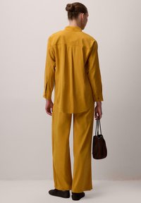 Žlutá košile s dlouhým rukávem, límcem a texturovanými detaily, spárovaná s odpovídajícími širokými kalhotami. Materiál vypadá lehký a splývavý.