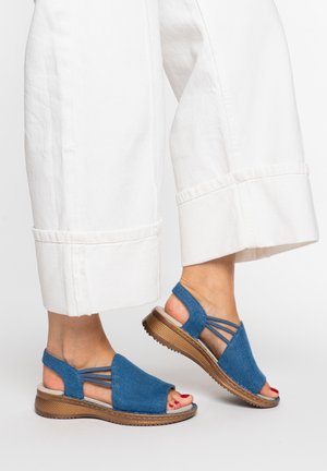 Stopy w niebieskich sandałach z odkrytymi palcami, z elastycznymi paskami i brązowymi podeszwami, w połączeniu z białymi, szerokimi, 7/8 spodniami.