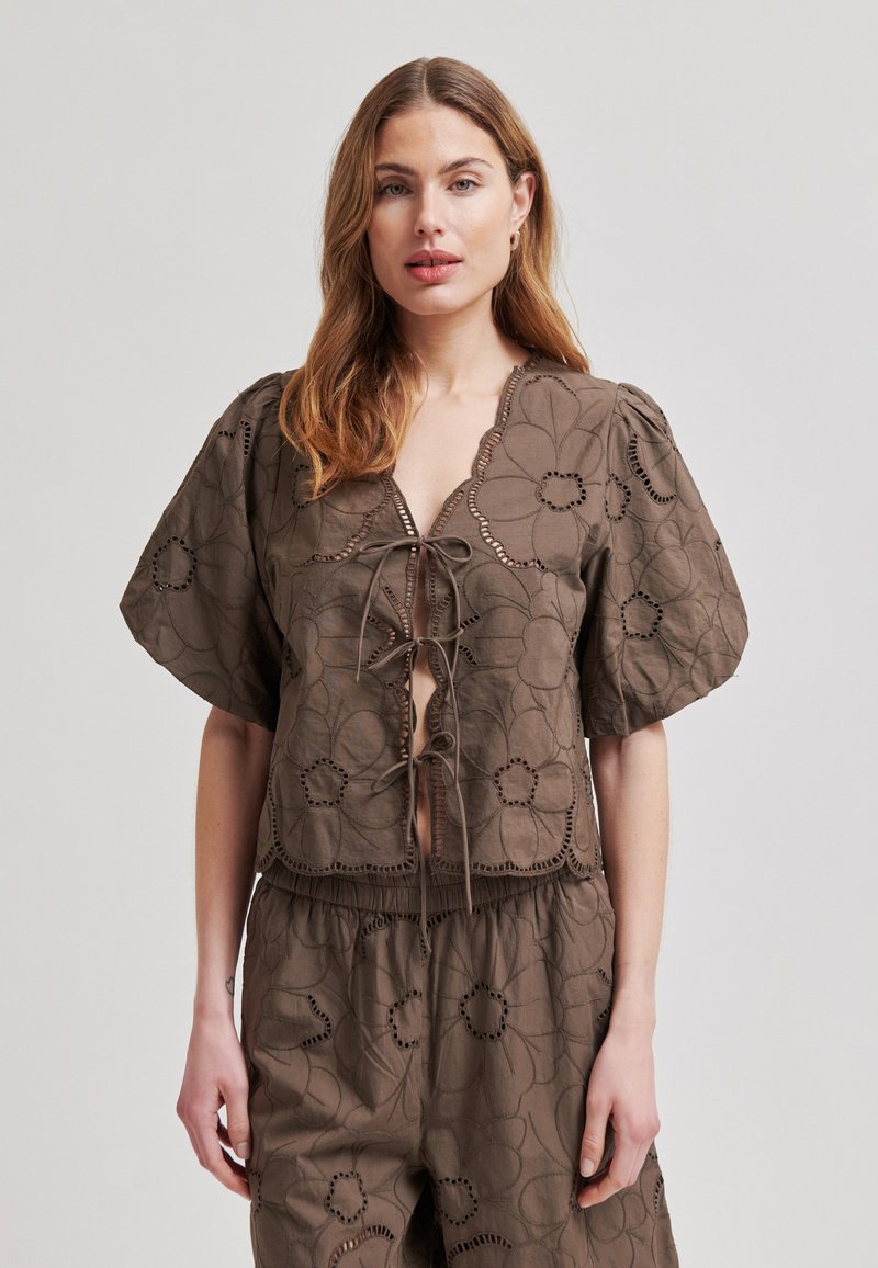 Femme portant un haut marron à fleurs brodées avec des manches bouffantes et un pantalon assorti, debout devant un fond clair uni.