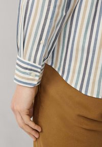 Camisa de botones a rayas en blanco, azul y beige. Presenta un cuello clásico, puños con botón y un dobladillo curvado. Se usa con pantalones marrones.