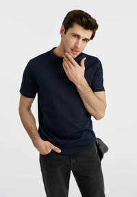 Granatowa koszulka z krótkim rękawem wykonana z gładkiego materiału, z okrągłym dekoltem i dopasowanym fasonem, noszona z ciemnymi dżinsami.
