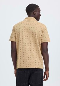 Polo à manches courtes en tissu beige avec un motif texturé de petits points noirs. Il présente un col classique et un ourlet droit.