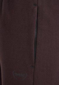 Dunkelbrauner Sweatshirtstoff mit schwarzer Reißverschlusstasche und dezentem Logodetail. Glatte Textur mit verstärkten Nähten an den Kanten.