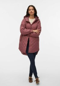 Ženska stoji z roki prekrižanimi, oblečena v vijolično odejasto kapuco, temne oprijete kavbojke, krem pulover in rjave čevlje na belem ozadju.