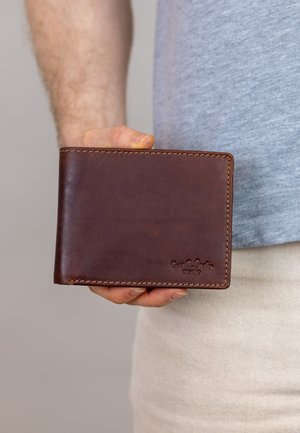 Bruine leren portemonnee met zichtbare stiksels en een geëmbossde logo, vastgehouden in een hand. De portemonnee heeft een gladde textuur en een compact ontwerp.