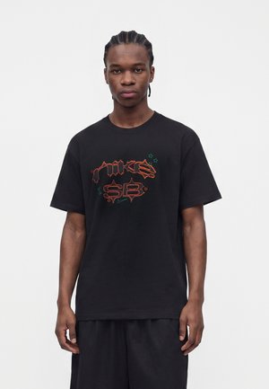 Młody mężczyzna z zaplecionymi włosami, ubrany w czarny t-shirt z neonowo pomarańczowo-zielonym logo „Nike SB” w stylu graffiti, stojący na jednolitym białym tle.
