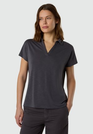 Donna con capelli castani di media lunghezza che indossa una polo grigia scura a maniche corte e pantaloni scuri abbinati, in piedi con una mano in tasca.