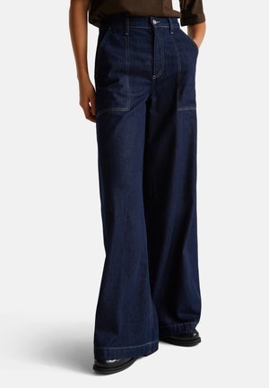 Persoon in donkerblauwe wijde denimjeans met voorzakken, zwarte schoenen en een bruine top met de handen in de zakken.