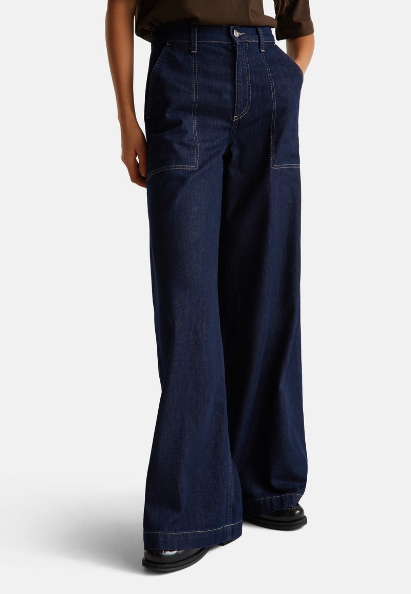 Široké tmavě modré džíny s výraznými předními kapsami a kontrastním prošíváním. Materiál má hladký, strukturovaný povrch.