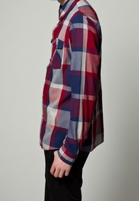 Person klädd i en långärmad skjorta med rutigt mönster i rött, vitt och blått, med knäppta manschetter och svarta byxor, som står på sidan mot en neutral bakgrund.