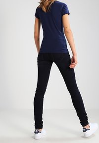 Mujer de pie, de espaldas, vistiendo una camiseta azul marino, jeans ajustados negros y zapatillas Adidas blancas sobre un fondo liso.