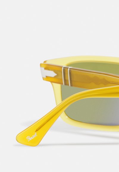 Ochelari de soare galbeni cu un cadru transparent galben și maro, lentile verzi și un brad texturat cu detalii ale logo-ului.