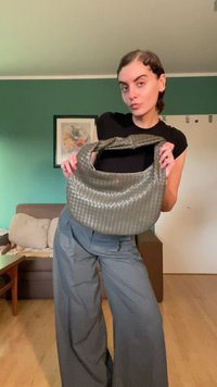 Borsa grigia intrecciata con forma semicircolare e un manico singolo; presenta una texture liscia e una sottile lucentezza, che si abbina a pantaloni grigi larghi.