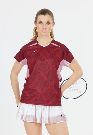 Kvinde badmintonspiller i vinrød sportstrøje og hvid nederdel, der holder ketcher bag ryggen og kigger fremad med neutralt udtryk.