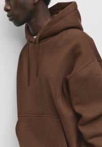 Brown hoodie with a relaxed fit, featuring a kangaroo pocket, drawstring hood, and soft texture. Accent stitching visible along seams.