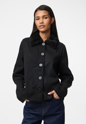 Veste noire raccourcie avec un col en fausse fourrure, dotée de grands boutons noirs, de manches longues et d'un tissu texturé. Le mannequin pose les mains dans les poches.