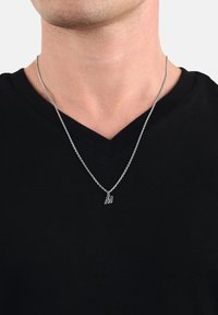 Silverkedja med ett "M"-buchstavsberlock bärs av en person klädd i en svart V-ringad tröja, visad från nacke till bröst.