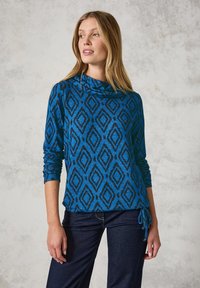Blauwe trui met zwart ruitpatroon, coltrui en lange mouwen; heeft een trekkoorddetail in de taille. Gekleed met donkere jeans.