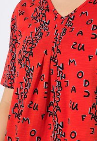 Top rojo de manga corta con escote en V y detalle de pliegue. La tela presenta un patrón caótico de letras y números en negro.