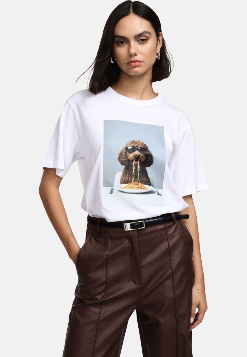 Maglietta bianca in cotone con una grafica di un cane con occhiali da sole, seduto a un tavolo con un piatto di spaghetti. Pantaloni in pelle marrone.