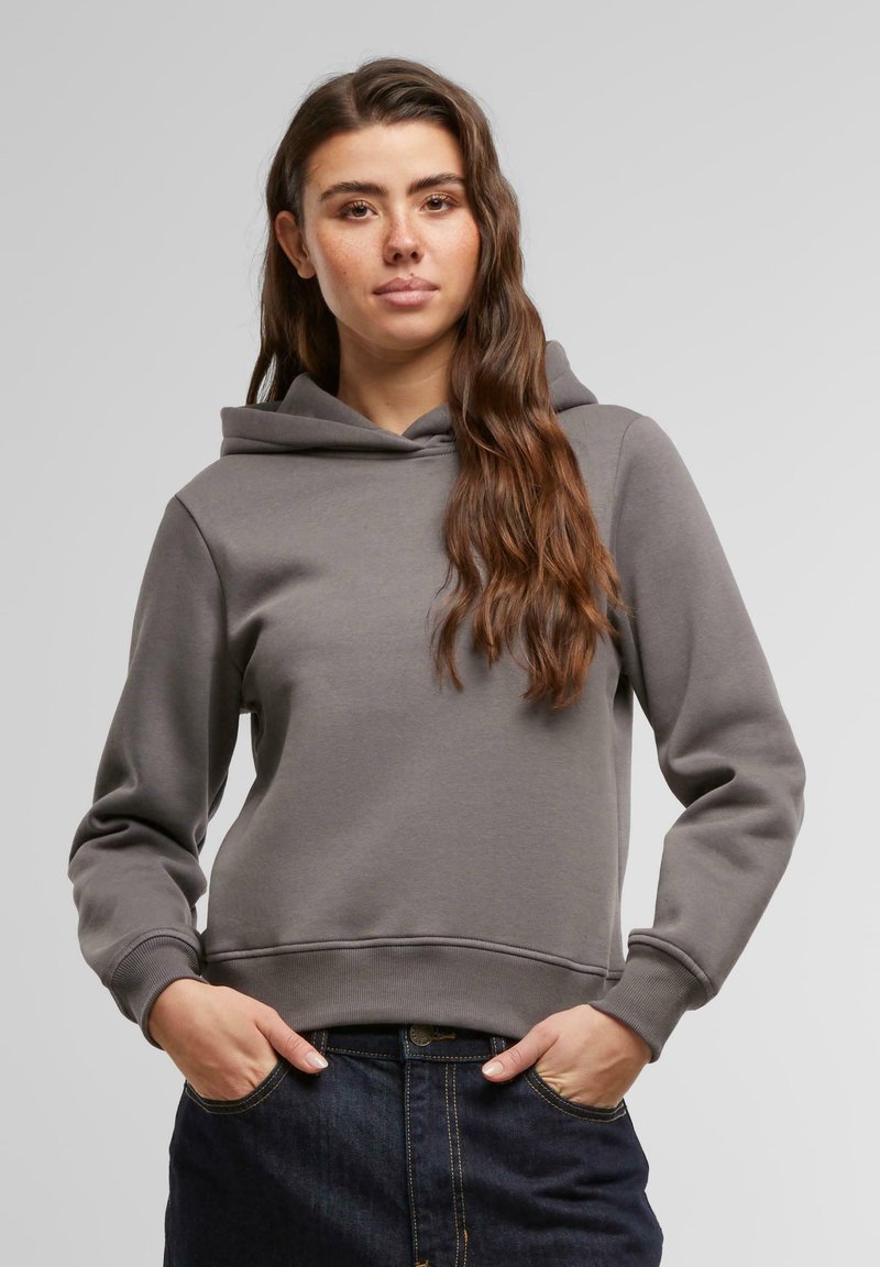 Hoodie gris avec une poche kangourou, texture douce, poignets et ourlet côtelés, et capuche à cordon. Porté par-dessus une jupe en jean foncée.