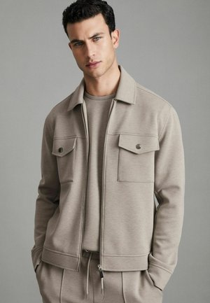 Beige zip-up jacket with two chest pockets, vertical stitching, and a smooth texture, paired with a matching crew-neck shirt.