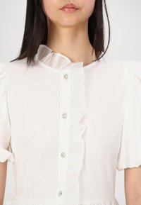 Vit klänning med volangkrage och korta puffärmar, som har dekorativa knappar framtill och ett mjukt, lätt tyg.