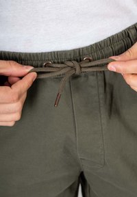 Pantalones verde oliva con cinturilla elástica y cordón. Presentan ojales de metal para el cordón y costuras laterales cosidas.