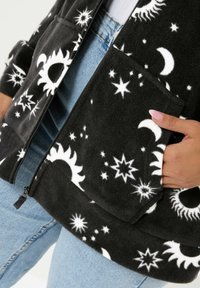 Mustan fleece-takkia, jossa on valkoisia taivaankappaleita, kuten tähtiä ja kuita, korostavia kuvioita. Varustettu täysipitkällä vetoketjulla ja etutaskuilla.