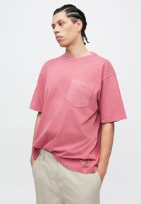 Ροζ φαρδύ μπλουζάκι με στρογγυλή λαιμόκοψη και τσέπη στο στήθος. Το ύφασμα φαίνεται μαλακό και ελαφρώς υφασμένο. Φορεμένο με ανοιχτό μπεζ σορτς.