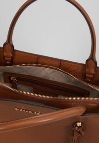 Brązowa skórzana torba na ramię o strukturalnym wykończeniu, z podwójnymi uchwytami i złotymi akcentami. Wnętrze zawiera kieszenie zamykane na zamek oraz otwarte, jasne podszycie.