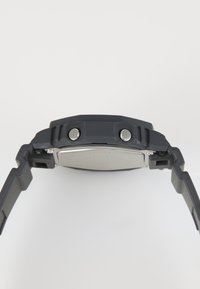 Montre en caoutchouc noir avec un boîtier épais, des accents argentés et un bracelet texturé. Comprend deux boutons sur le côté et un dos métallique lisse.