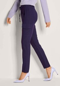 Vrouw in een marineblauwe cargobroek en lichtpaarse hoge hakken, gecombineerd met een lichtpaarse trui, die loopt tegen een beige muur als achtergrond.