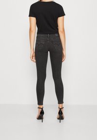 Vaqueros negros de cintura alta y corte ajustado con un tejido texturizado, dos bolsillos traseros con costura en forma de corazón, combinados con una blusa negra y tacones altos.