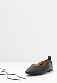 Chaussure plate de ballet noire texturée avec un languette en cuir au talon et un lacet noir détaché sur une surface blanche.