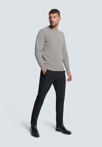 Lichtgrijze geribde trui met lange mouwen, gecombineerd met slanke zwarte broek en enkellaarsjes. Model staat met handen in de zakken.