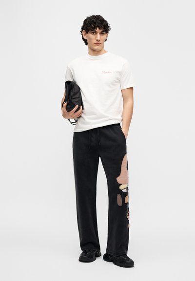 Άσπρο βαμβακερό t-shirt με κόκκινο κεντημένο λογότυπο, συνδυασμένο με μαύρο παντελόνι φόρμας με πολύχρωμες υφασμάτινες ταινίες και πλαϊνές τσέπες, φορεμένο με μαύρα παπούτσια.