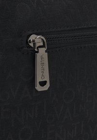 Kovový tahák na zip s vyrytým nápisem "VALENTINO" na černé tkanině vzorované opakujícím se textem "VALENTINO".