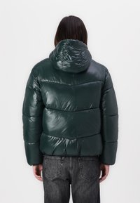 Chaqueta acolchada verde tipo abrigo con un acabado brillante, que incluye capucha y secciones acolchadas distintas. Combinada con jeans oscuros y un corte relajado.