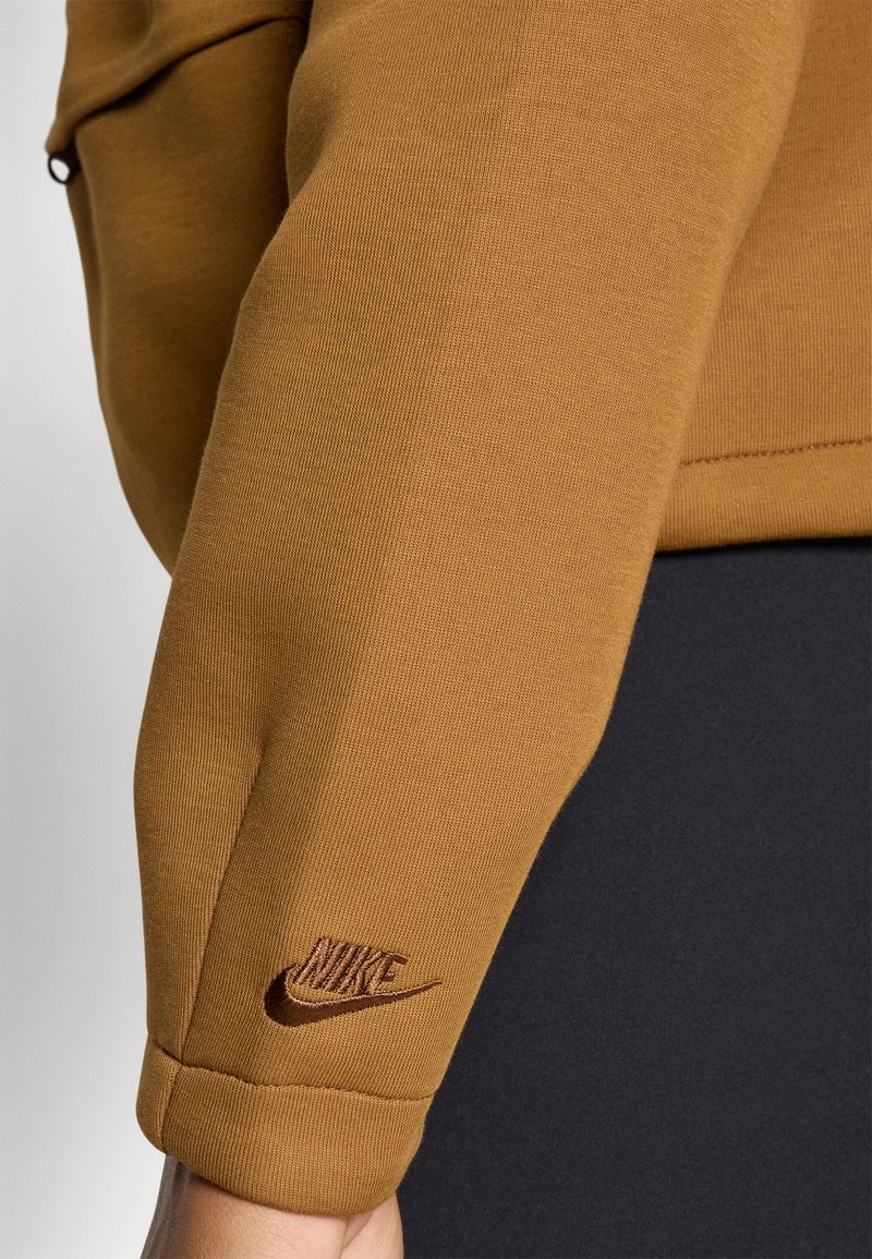 Brauner Pullover mit strukturierter Oberfläche, ausgestattet mit einem Bündchenärmel und einem aufgenähten braunen Nike-Logo am Handgelenk. Schwarzer Stoff darunter.