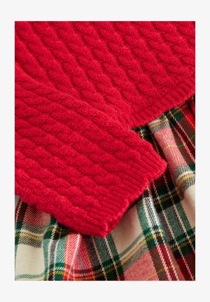 Roter Strickpullover mit einem Kabelmuster, der über einem karierter Rock mit grünen, roten und gelben Linien auf einem elfenbeinfarbenen Hintergrund getragen wird.