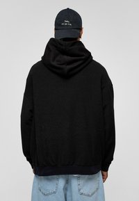 Hoodie noir oversize avec une grande capuche et un ourlet côtelé. Porté avec un jean baggy bleu clair et une casquette sombre avec du texte blanc.