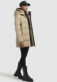 Beige pufferjack met capuchon, voorzien van een ritssluiting en een contrasterende voering, gecombineerd met zwarte leggings en veterschoenen.