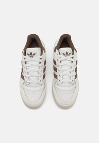 White leather sneakers with brown textile accents, featuring three side stripes, round toe, perforated detailing, and white laces.