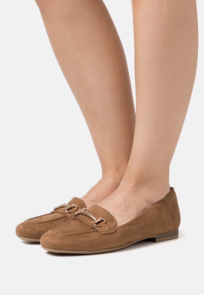 Paire de mocassins plats en daim marron avec détail boucle dorée portés pieds nus sur fond blanc.