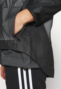 Main placée dans la poche zippée d'une veste noire texturée portée sur un pantalon noir à rayures verticales blanches.