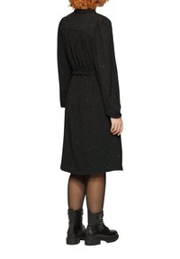 Schwarzes Kleid mit langen Ärmeln, figurnaher Taille und Rückenlänge bis zur Mitte der Waden. Der Stoff hat einen dezenten Glitzer und ein gefälteltes Rückendetail. Kombiniert mit schwarzen Kampfstiefeln.
