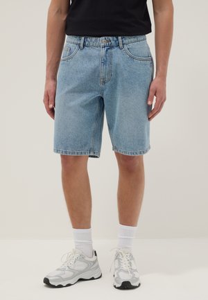 Parte inferiore del corpo che indossa pantaloncini di jeans azzurri chiari, calzini bianchi e scarpe sportive bianche, in piedi davanti a uno sfondo semplice.