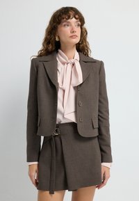 Brązowa marynarka dopasowana z złotymi guzikami, w połączeniu z jasnoróżową bluzką z kokardą i dopasowaną spódnicą o wysokim stanie. Gładka tekstura.